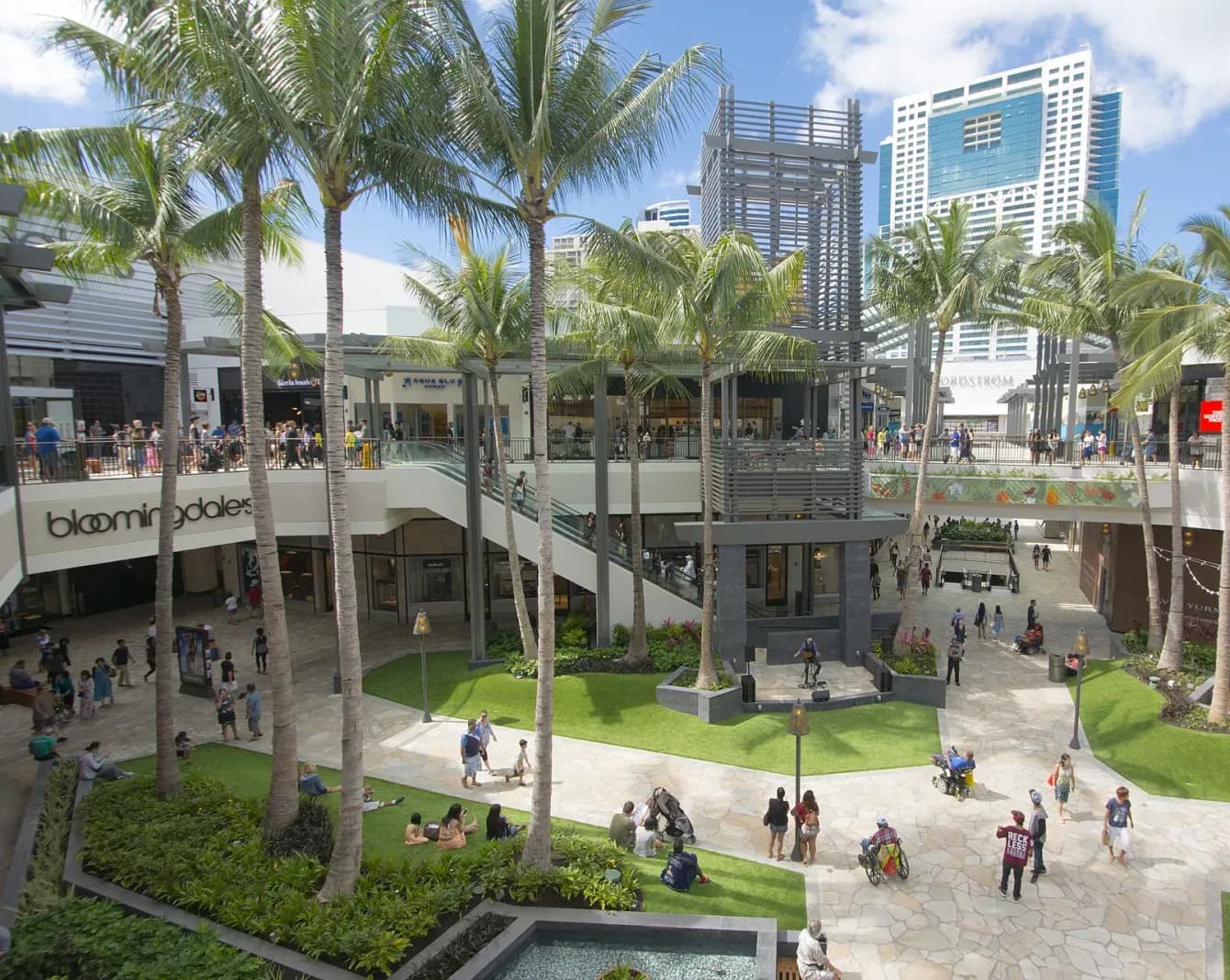 Khoảng sân ngoài trời của Ala Moana Center với hàng dừa, thảm cỏ, khách đi dạo và các tòa nhà cao phía sau (ảnh chụp năm 2015)