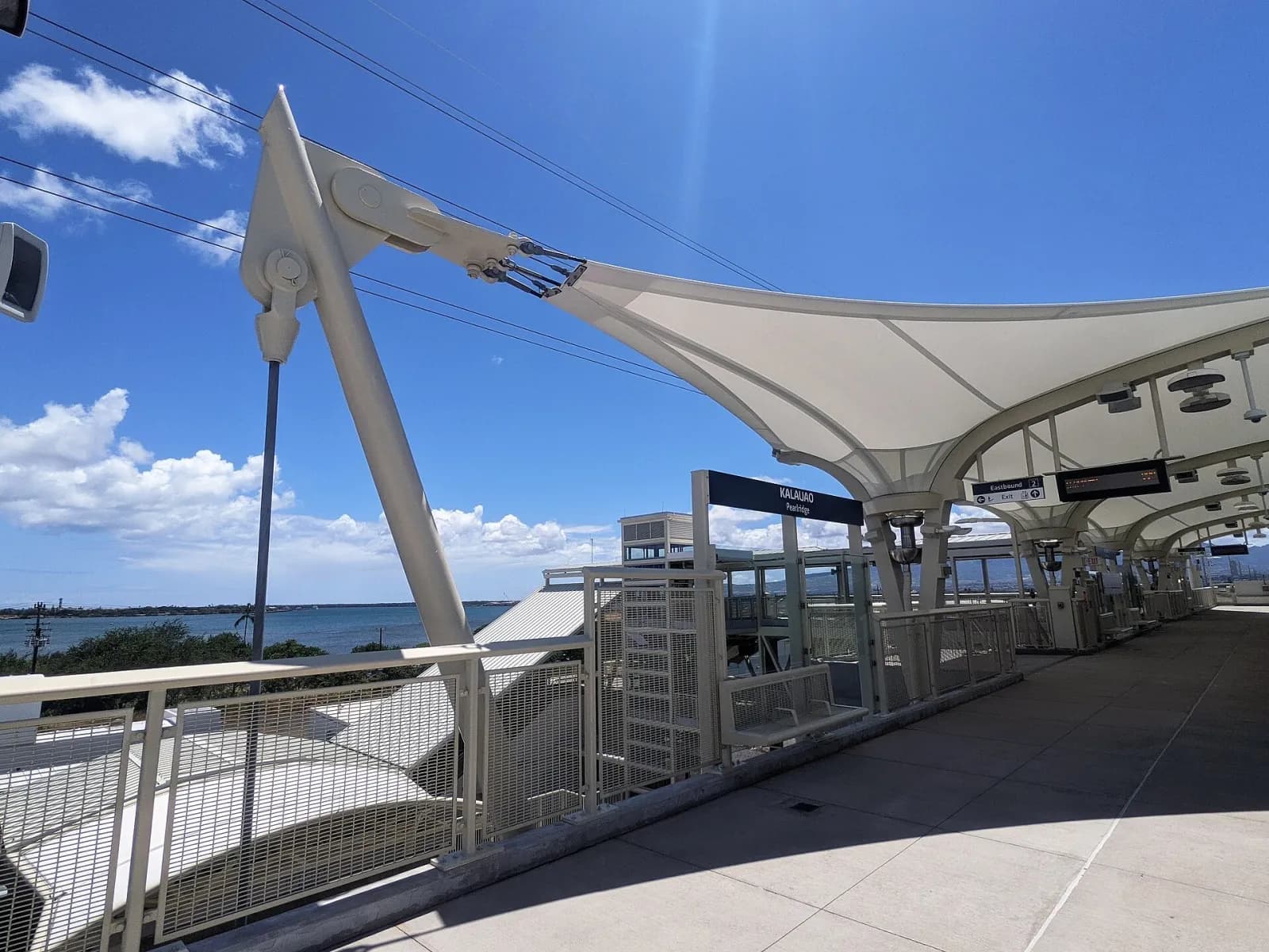 Sân ga trạm Skyline Kalauao (Pearlridge) với mái che màu trắng, bảng tên trạm và biển phía xa, không phải bên trong trung tâm mua sắm
